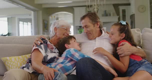 Πρόσοψη των Καυκάσιων παππούδων που κάθονται σε έναν καναπέ στο σαλόνι αγκαλιασμένοι με τον εγγονό και την εγγονή τους, κάνοντας γκριμάτσες και χαμογελώντας ο ένας στον άλλο, αργή κίνηση - Πλάνα, βίντεο