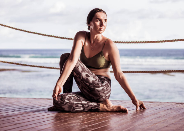 Young caucasian woman practicing yoga, sitting on wooden floor in Ardha Matsyendrasana. Half spinal twist pose. Ocean background. Zen life. Yoga retreat in Bali, Indonesia - Photo, Image