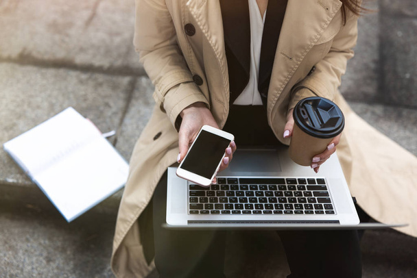 zakenvrouw houdt laptop op haar knieën met kopje koffie en smartphone in handen zitten op de trap in de buurt van kantoor tijdens lunchpauze, multitasking concept. - Foto, afbeelding
