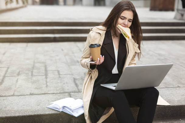 young beautiful businesswoman with sandwich in her mouth holding hot coffee in paper cup works in her laptop sitting on stairs outside office , multitasking concept. - Foto, imagen