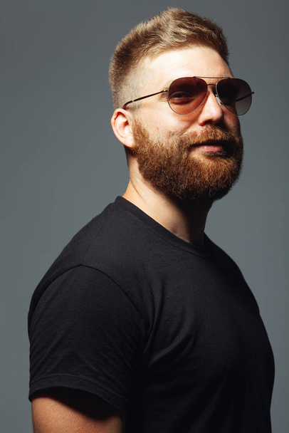 Eyewear concept. Fabulous at any age. Portrait of charismatic muscular 30-year-old man standing over dark gray background. Perfect haircut. Rocker, biker, hipster style. Studio shot - Foto, Bild