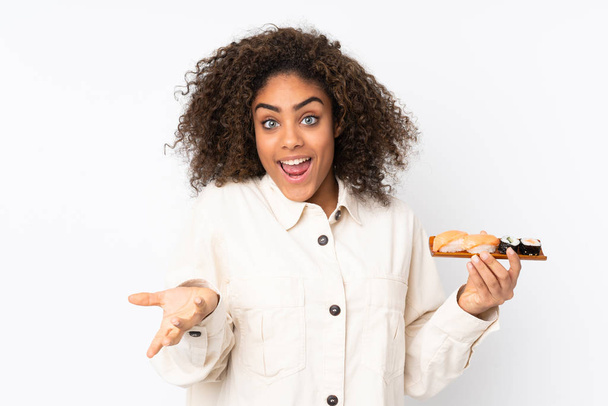 Молодая афроамериканка держит суши на белом фоне с шокированным выражением лица
 - Фото, изображение
