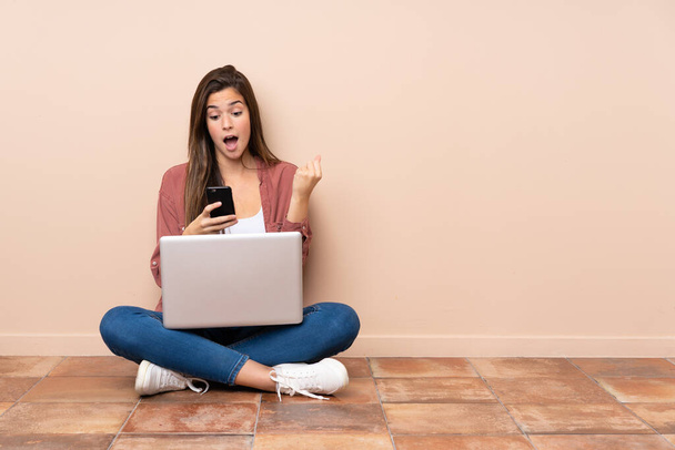 Adolescente étudiante assise sur le sol avec un ordinateur portable surprise et envoyant un message - Photo, image