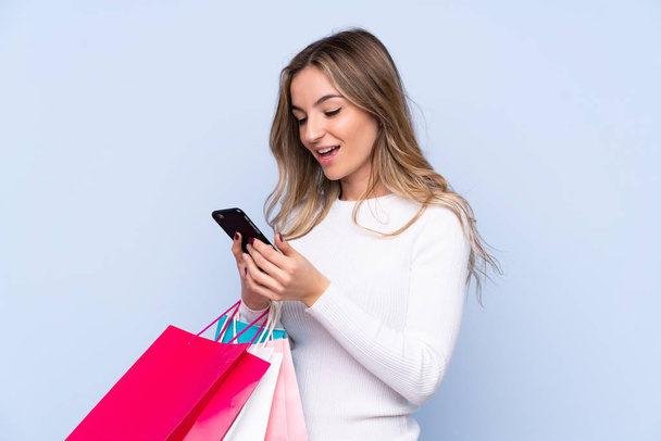 孤立した青の背景の上に若い女性ショッピングバッグを保持し、友人に彼女の携帯電話でメッセージを書く - 写真・画像