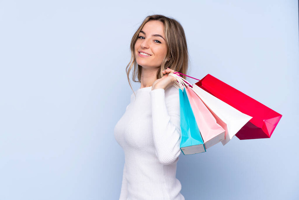 Junge Frau vor isoliertem blauen Hintergrund hält Einkaufstüten in der Hand und lächelt - Foto, Bild