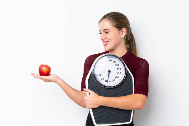 Teenager dívka s váhou nad izolované bílé pozadí drží vážicí stroj při pohledu na jablko - Fotografie, Obrázek