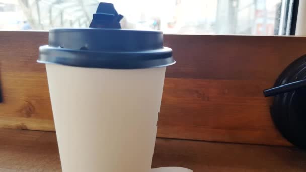 Er staat een witte papieren kop koffie op een houten tafel in het café, en door het glas zie je de straat met een wazige focus - Video