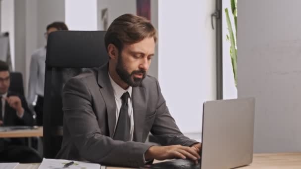 Serious businessman in formal suit using laptop computer and writing something in document while sitting by the table in office - Séquence, vidéo