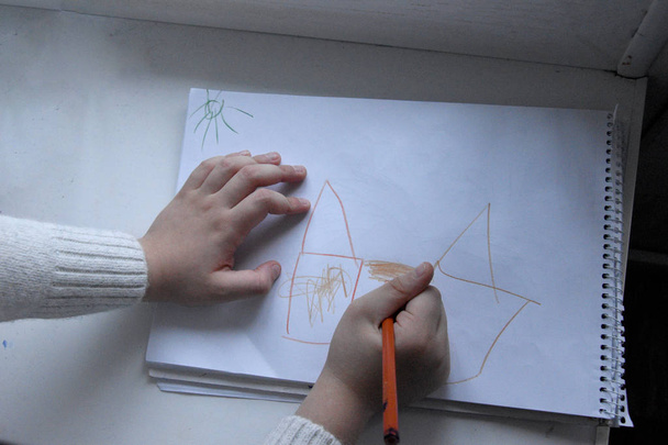 De jongens houden een potlood vast, het kind tekent meerdere tekeningen, een huis, een gezin. Kindertekeningen van het gezin en thuis. - Foto, afbeelding