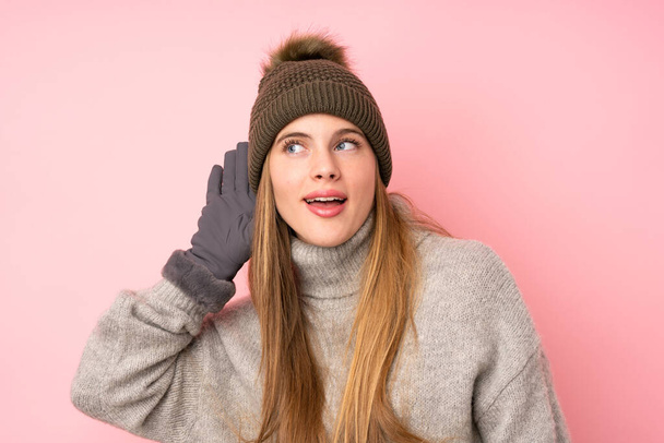 Jeune adolescente avec chapeau d'hiver sur fond rose isolé écoutant quelque chose
 - Photo, image