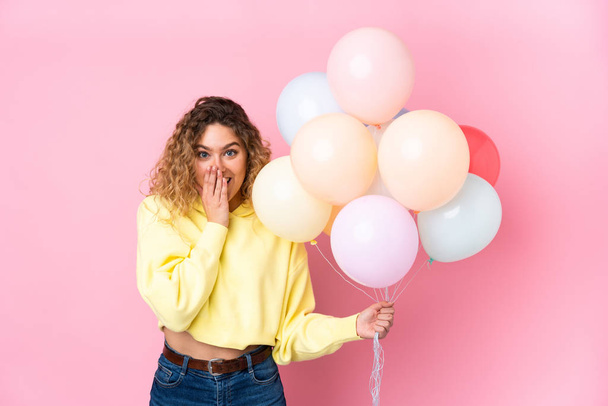 Young blonde woman with curly hair catching many balloons isolated on pink background with surprise facial expression - Photo, Image