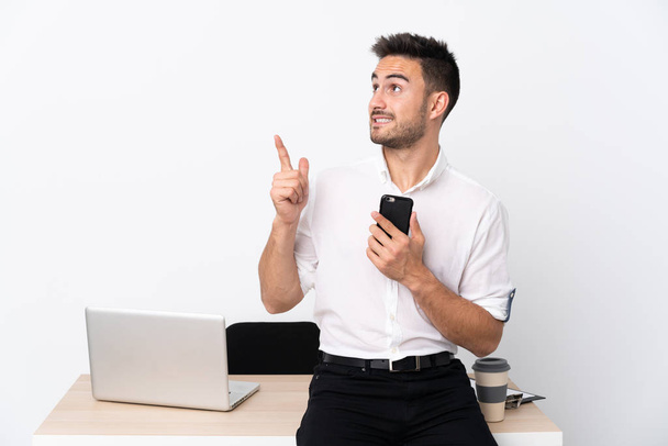 Молодой бизнесмен с мобильным телефоном на рабочем месте указывает указательным пальцем отличная идея
 - Фото, изображение