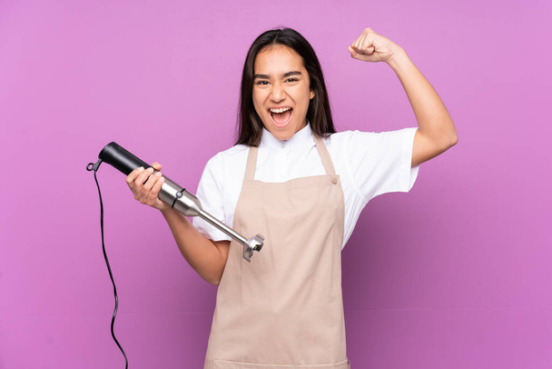 Indian woman using hand blender isolated on purple background celebrating a victory - Photo, Image