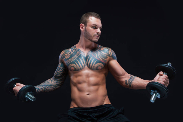 Tattooed strong muscular athletic man lifting up dumbbells on black background - Fotó, kép