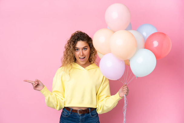 Jovem loira com cabelos cacheados pegando muitos balões isolados no fundo rosa surpreso e apontando o dedo para o lado
 - Foto, Imagem