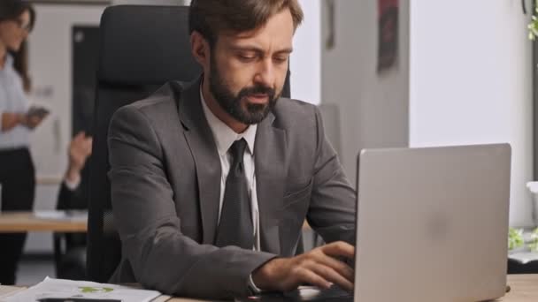 Serious businessman in formal suit using laptop computer and relaxing after that while sitting by the table in office - Materiał filmowy, wideo