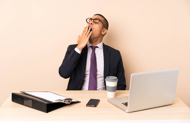 Молодий бізнесмен в своєму офісі з ноутбуком та іншими документами, що позіхають і покривають широкий відкритий рот рукою
 - Фото, зображення