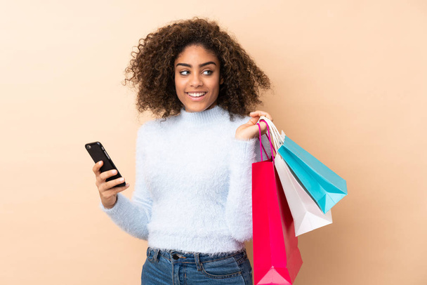 Young African American woman isolated on beige background holding shopping bags and a mobile phone - Fotó, kép