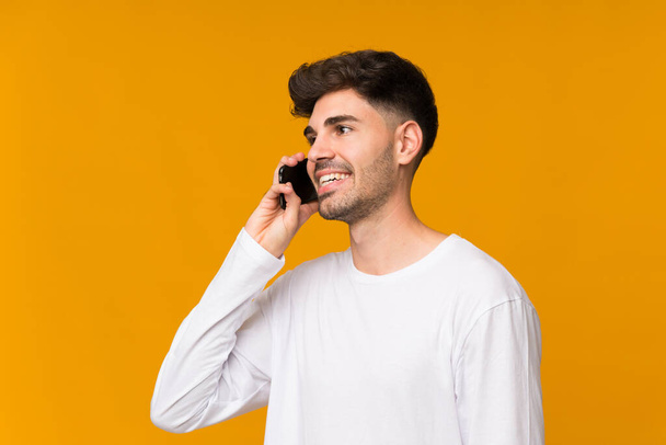 Молодой человек на изолированном оранжевом фоне поддерживает разговор с мобильным телефоном
 - Фото, изображение