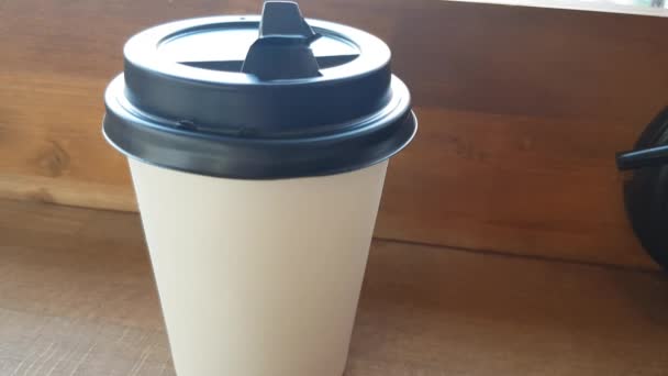 Υπάρχει ένα λευκό χαρτί Κύπελλο του καφέ σε ένα ξύλινο τραπέζι στο καφέ, και μέσα από το γυαλί μπορείτε να δείτε το δρόμο με μια θολή εστίαση - Πλάνα, βίντεο
