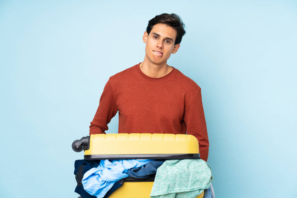 Homem com uma mala cheia de roupas sobre fundo azul isolado com dúvidas e com expressão facial confusa
 - Foto, Imagem