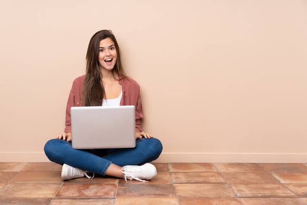 Adolescente estudiante chica sentada en el suelo con un ordenador portátil con la expresión facial sorpresa
 - Foto, imagen