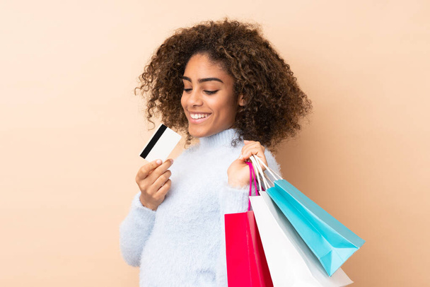Joven mujer afroamericana aislada sobre fondo beige sosteniendo bolsas de compras y una tarjeta de crédito - Foto, imagen