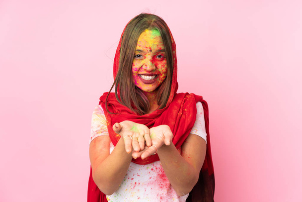 Young Indian woman with colorful holi powders on her face isolated on pink background holding copyspace imaginary on the palm to insert an ad - Photo, Image