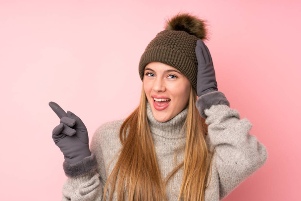 Молодая девушка-подросток в зимней шляпе на изолированном розовом фоне удивлена и указывает пальцем в сторону
 - Фото, изображение