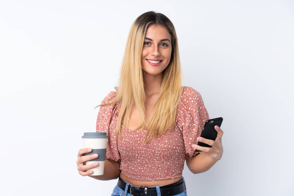 Young Uruguayan woman over isolated white background holding coffee to take away and a mobile - Foto, Imagen