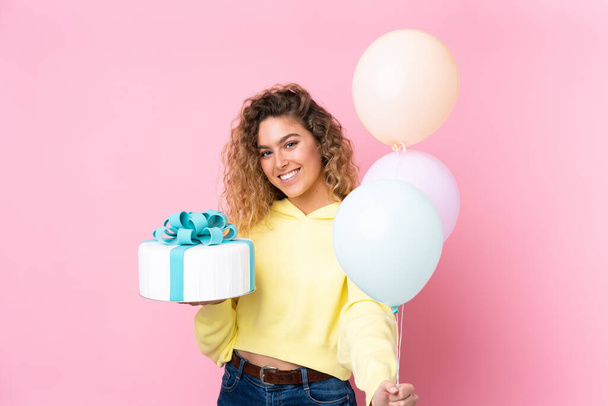 Young blonde woman with curly hair catching many balloons and holding a big cake isolated on pink background - Фото, изображение