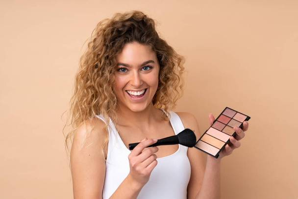 Young blonde woman with curly hair isolated on beige background with makeup palette and happy - Zdjęcie, obraz