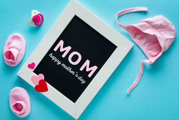 Vista superior de pizarra con letras de mamá cerca de ropa de bebé rosa y chupete sobre fondo azul
 - Foto, Imagen