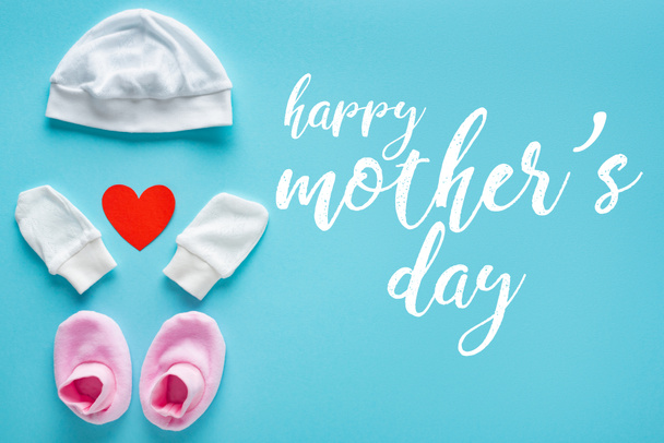Draufsicht auf Baby-Fäustlinge, Booties und Hut mit Papierherz auf blauem Hintergrund, glücklicher Muttertag Illustration - Foto, Bild