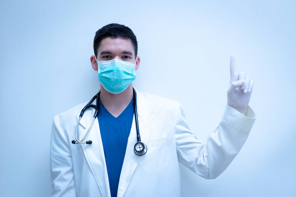 Un joven médico guapo cubriendo la cara con una máscara azucarada, vistiendo bata blanca uniforme y estetoscopio, apuntando con la mano izquierda hacia arriba
 - Foto, Imagen