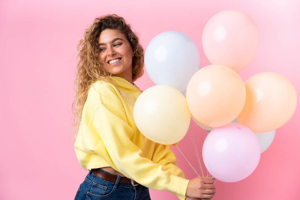 Young blonde woman with curly hair catching many balloons isolated on pink background - Fotografie, Obrázek