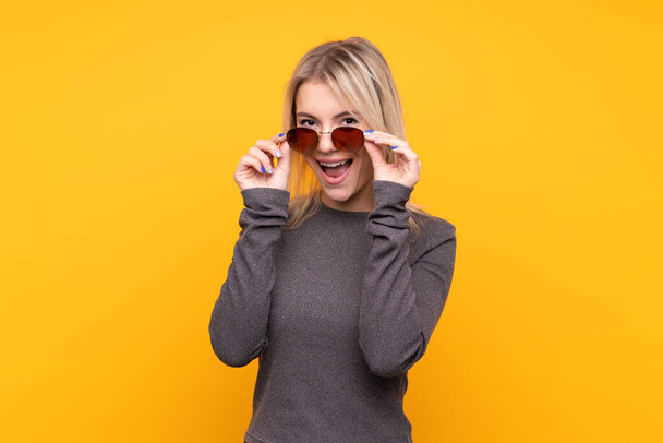 Giovane donna bionda su sfondo giallo isolato con occhiali e sorpreso
 - Foto, immagini