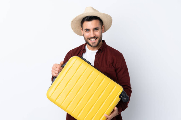 Jovem bonito homem sobre isolado fundo branco em férias com mala de viagem e um chapéu
 - Foto, Imagem