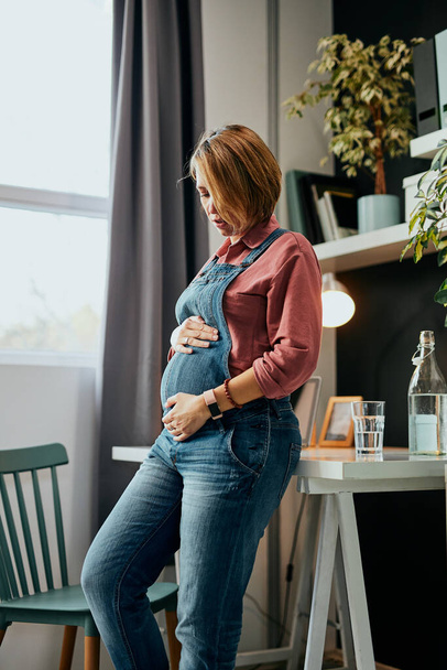 Proud gorgeous caucasian future mother leaning on the desk in her home office and touching her belly. - Photo, Image