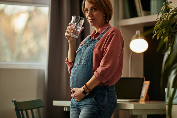Mujer embarazada de pie en su oficina en casa, tocando el vientre y bebiendo agua dulce
. - Foto, imagen
