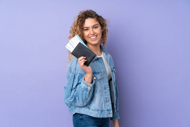 Mujer rubia joven con el pelo rizado aislado sobre fondo púrpura feliz en vacaciones con pasaporte y billetes de avión - Foto, Imagen