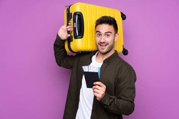 Young handsome man over isolated purple background in vacation with suitcase and passport - Photo, image