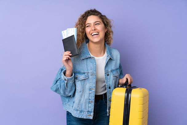 Young blonde woman with curly hair isolated on purple background in vacation with suitcase and passport - Foto, Imagem