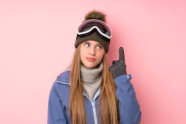 Snowboard gözlüklü kayakçı kız izole edilmiş pembe arka planda işaret parmağıyla işaret etmek harika bir fikir. - Fotoğraf, Görsel