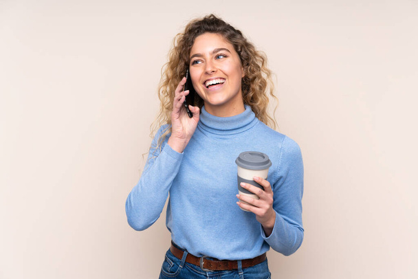 Fiatal szőke nő göndör haj visel garbó pulóver elszigetelt bézs háttér gazdaság kávé elvenni, és egy mobil - Fotó, kép