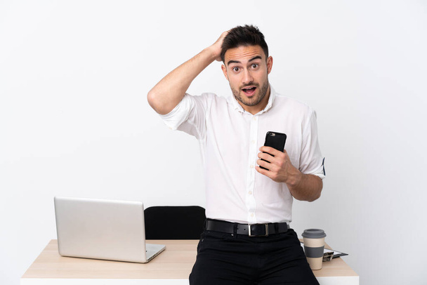 Jonge zakenman met een mobiele telefoon op een werkplek met verrassende gezichtsuitdrukking - Foto, afbeelding