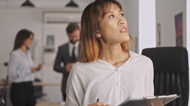 Pensive asian businesswoman holding clipboard and looking away while standing in office - Video