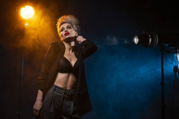 A beautiful blonde girl with an elegant hairstyle and large breasts, wearing a bra, trousers and a blazer, artistically poses in the rays of spotlights in the smoke. Cinematic, art, commercial design - Φωτογραφία, εικόνα