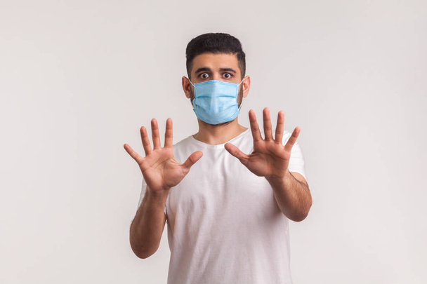 Alarming scared panicking man in hygienic mask gesturing stop, afraid of coronavirus infection, respiratory illnesses such as flu, 2019-nCoV, ebola. indoor studio shot isolated on white background - 写真・画像