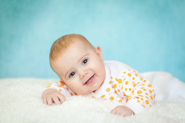 Happy excited 4 months infant baby girl lying on the bed with white fur. Cute girl wearing white clothes with orange polka dot on bright blue background. Textile and bedding for kids - Photo, image